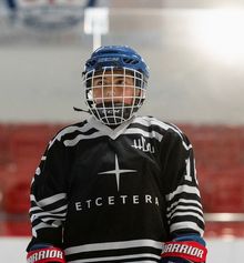 A kid in a black and white Etcetera hockey jersey standing with hockey boards behind him