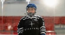 A kid in a black and white Etcetera hockey jersey standing with hockey boards behind him